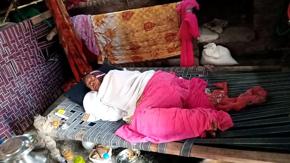An older woman in India rests under a blanket on a woven cot in her home.