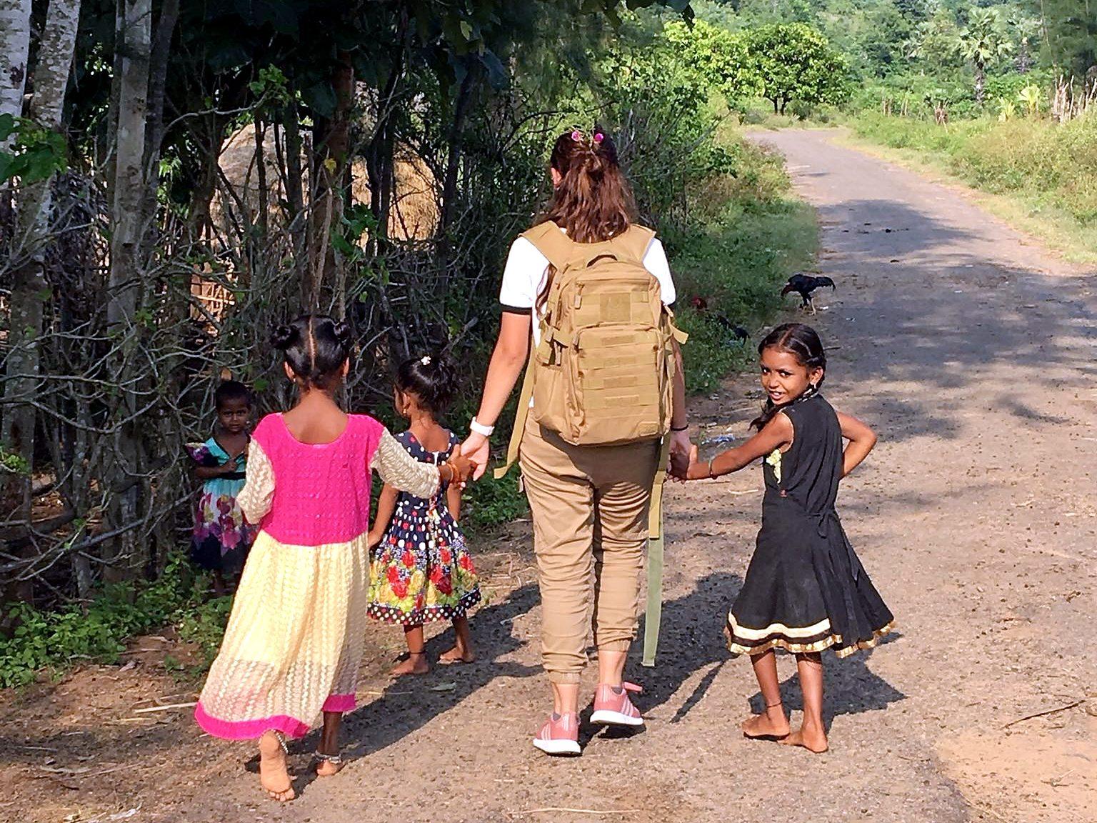 A woman with a backpack walks hand in hand with village children along a country road in India.