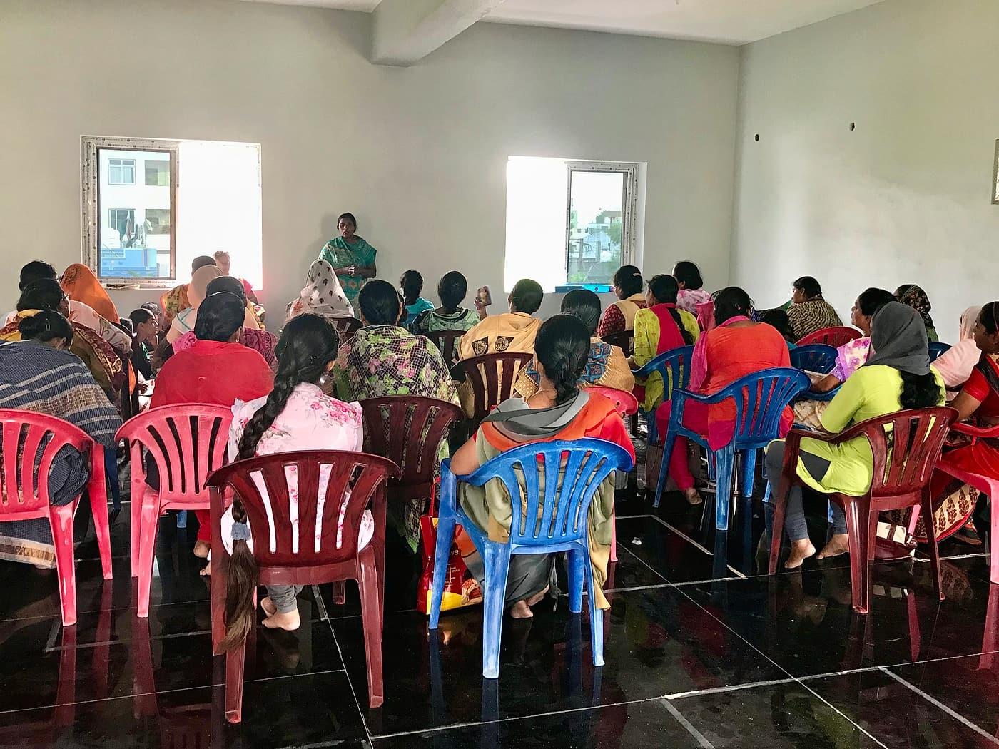 Women in bright saris sit on plastic chairs in a hall in India, listening to a woman leading a workshop at the front.