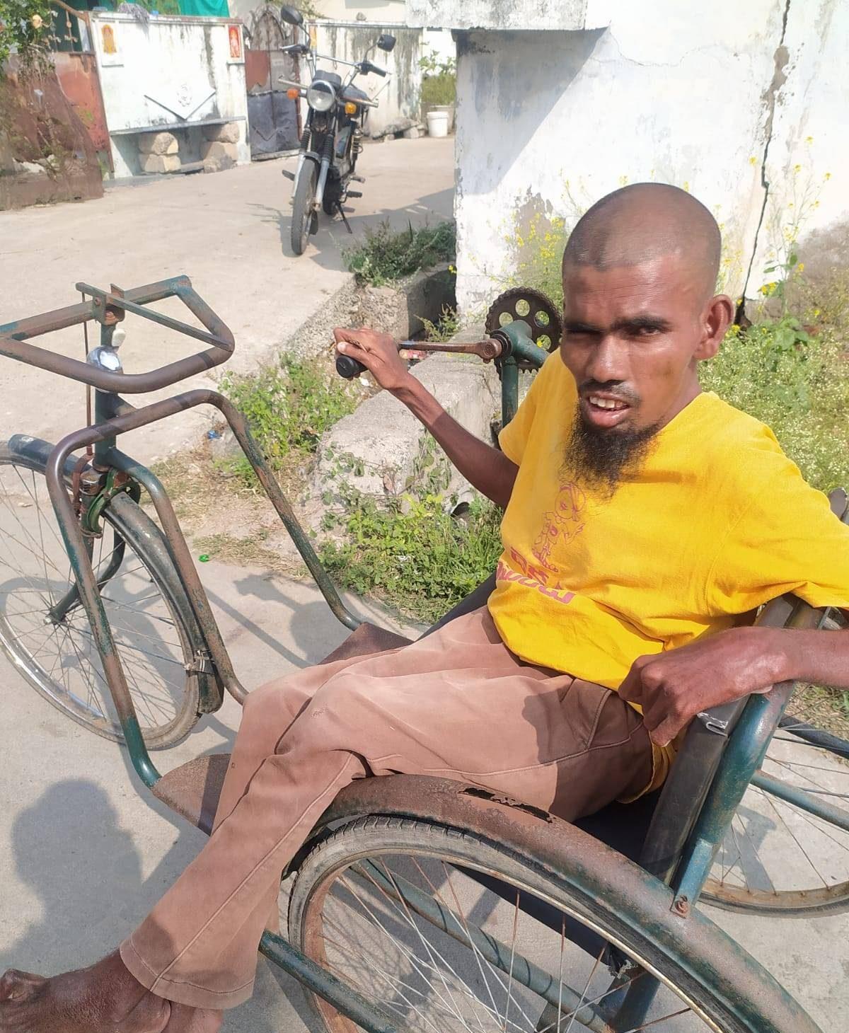 A man in a yellow shirt sits in a hand-cranked tricycle wheelchair on a village street in India.