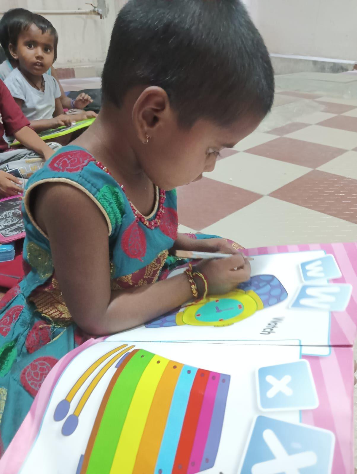 A young girl in India colours a picture during a lesson, other children working on the floor behind her.