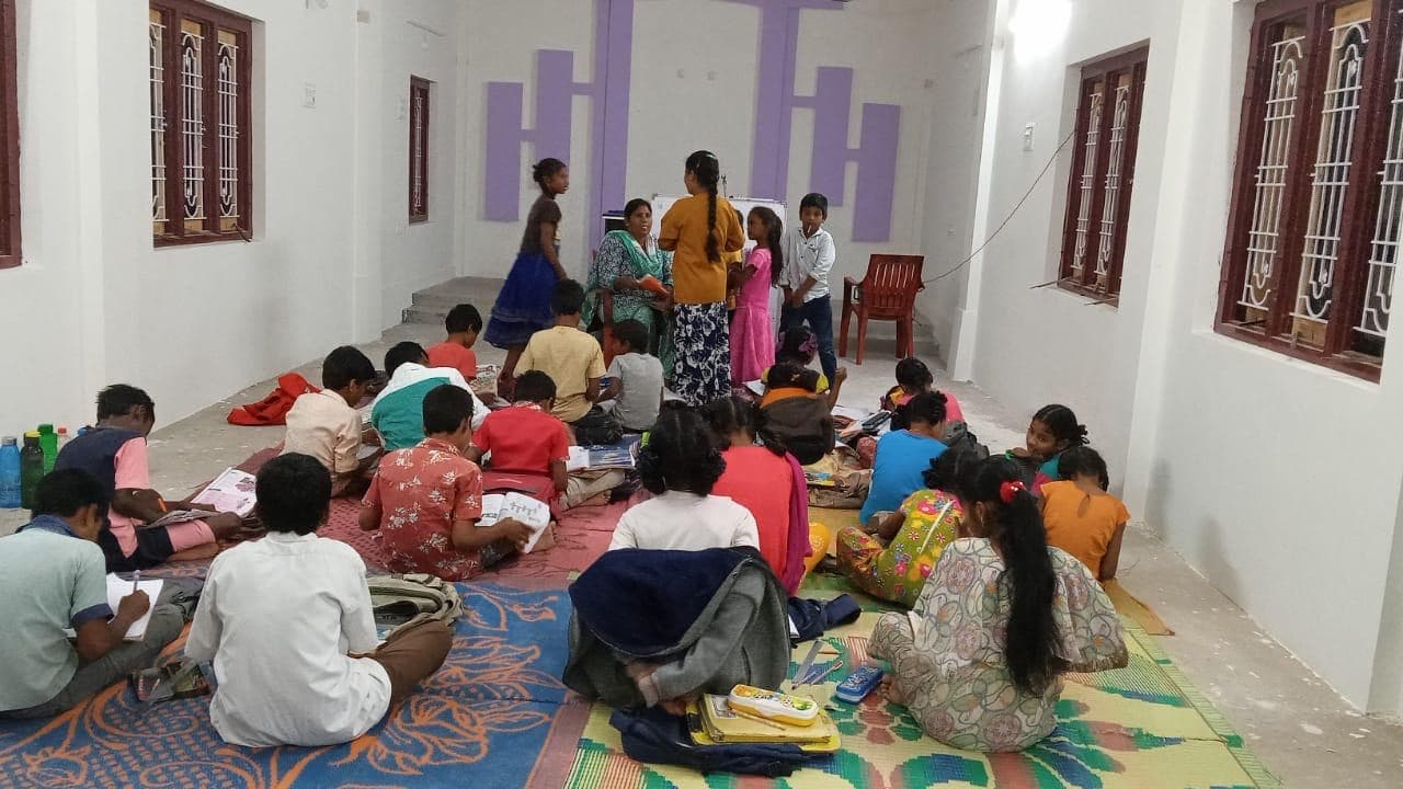 Children study on colourful mats in a bright classroom in India while a teacher works with a small group.